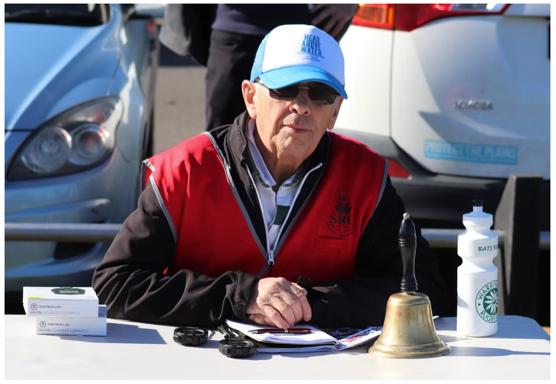Phil Jackson seated outdoors wearing sunglasses and a blue cap at community event
