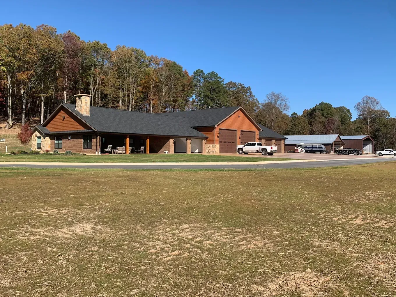 A single-story house with a covered porch, garage, and outbuildings; brown and tan, set on a grassy field with trees in the background.
