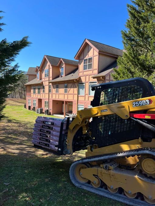 A yellow skid steer carrying purple insulation near a row of tan townhouses on a sunny day.