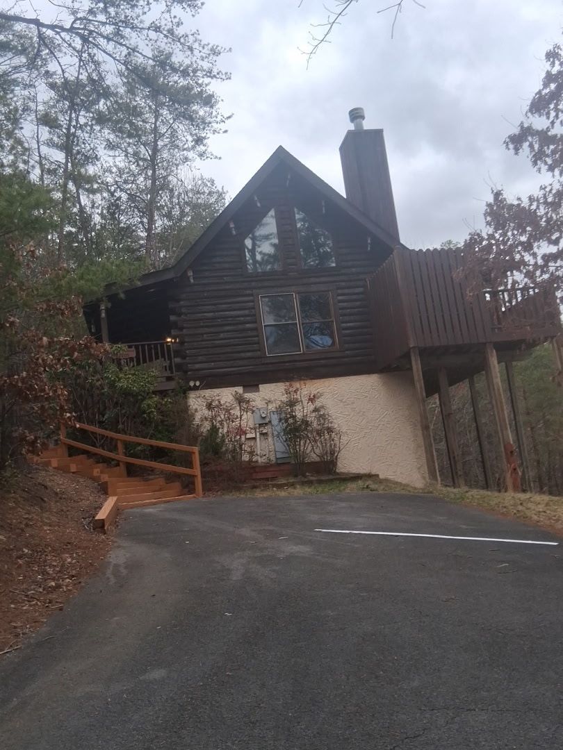 A rustic two-story log cabin sits on a hillside with a stone-colored foundation, chimney, and an asphalt driveway.