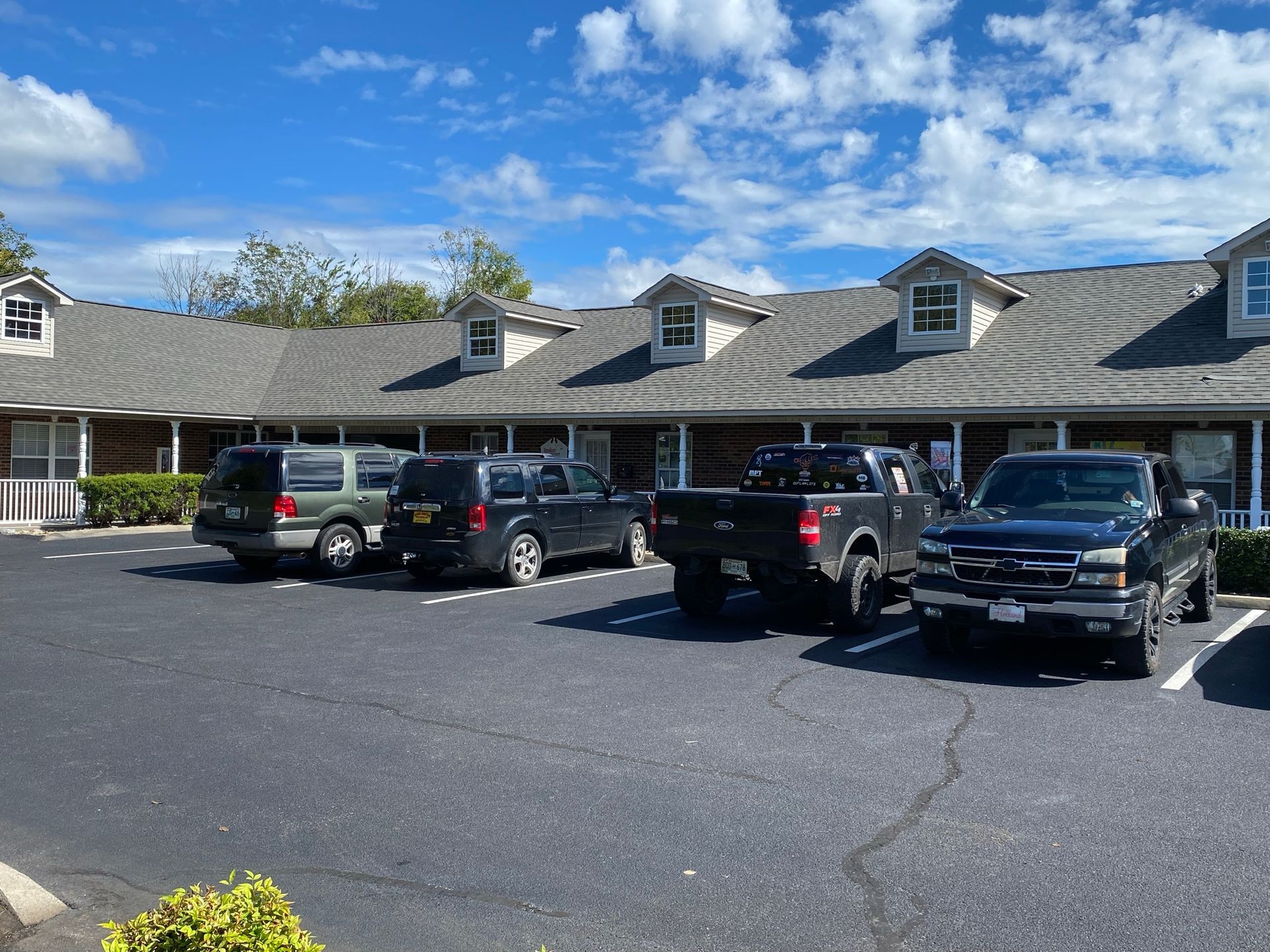 A low, one-story commercial building with grey shingles and dormer windows, featuring a parking lot with four vehicles.