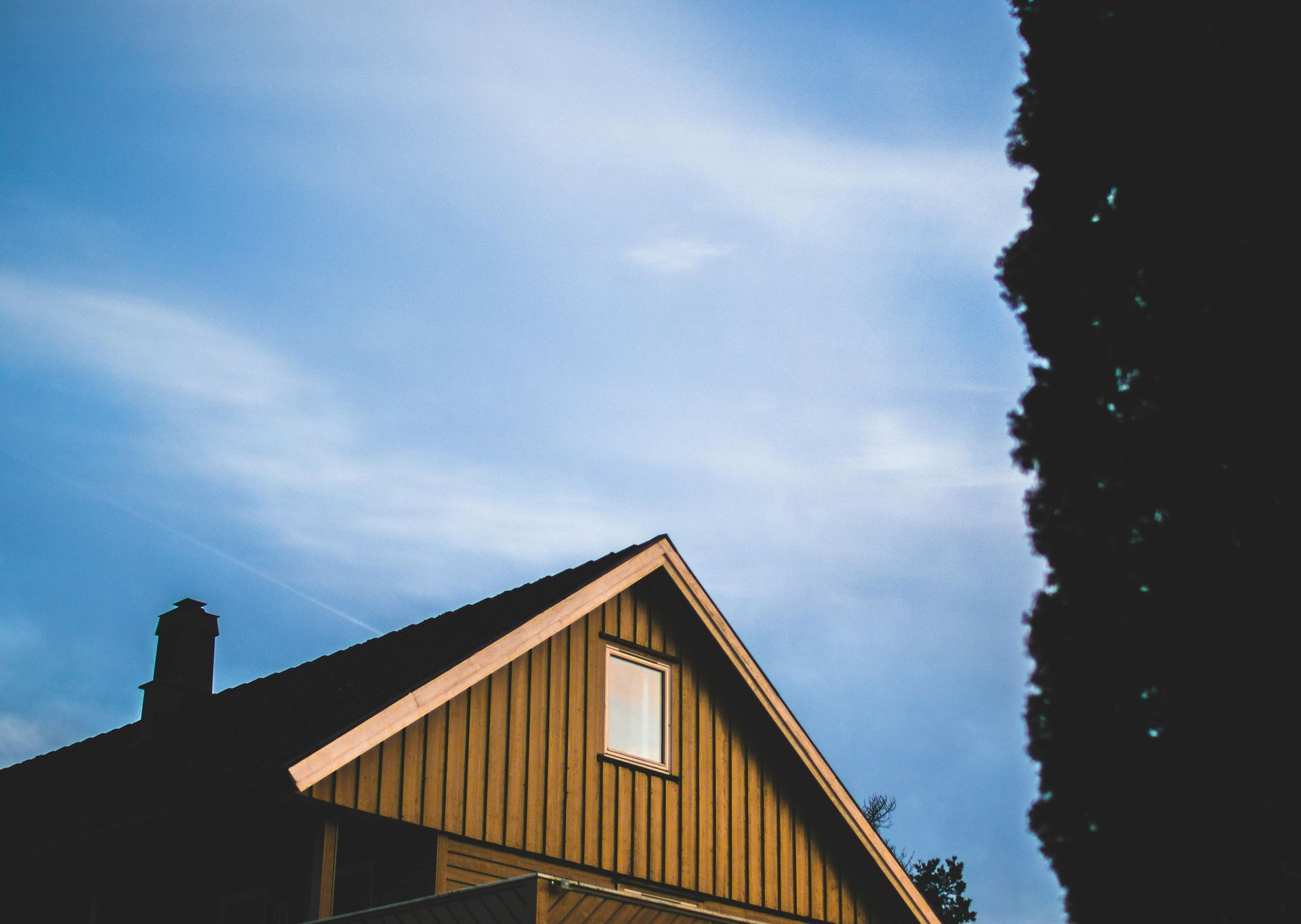 Wooden house gable against a blue sky, with a tall, dark tree on the right side.