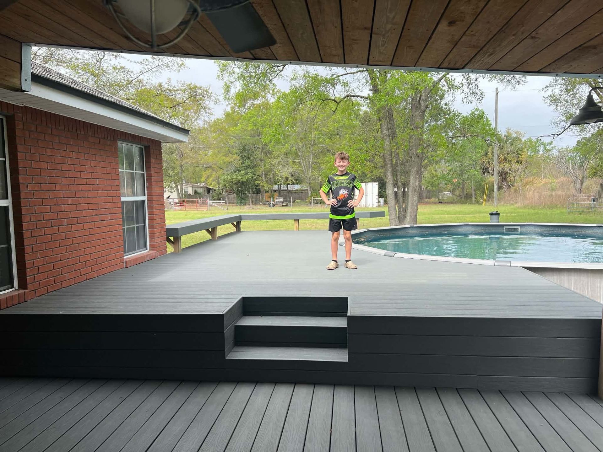 Person standing on a backyard deck beside a pool, with trees and a brick house nearby.