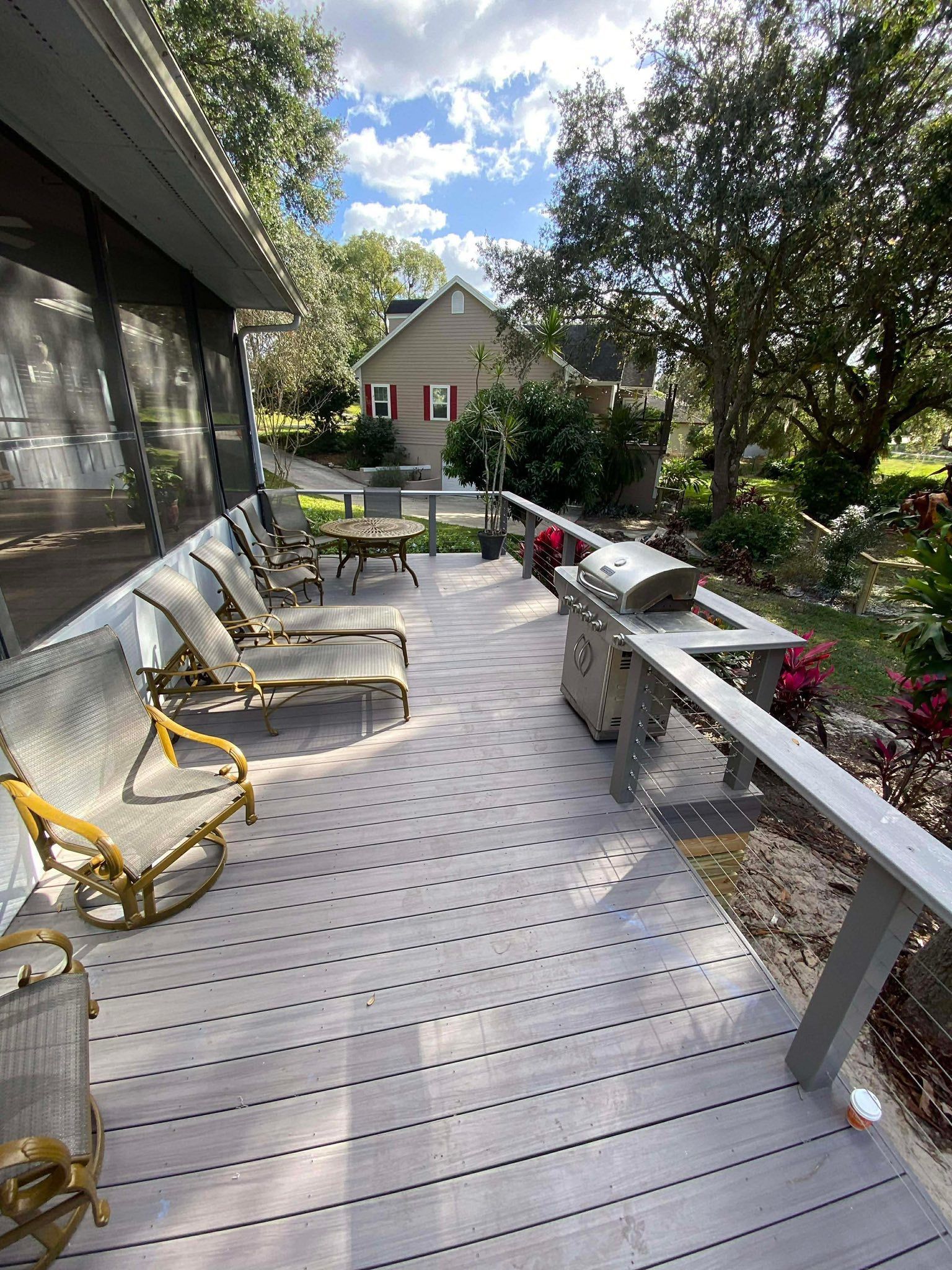 Outdoor deck with chairs and table, white railing, potted flowers, and trees in a sunny backyard