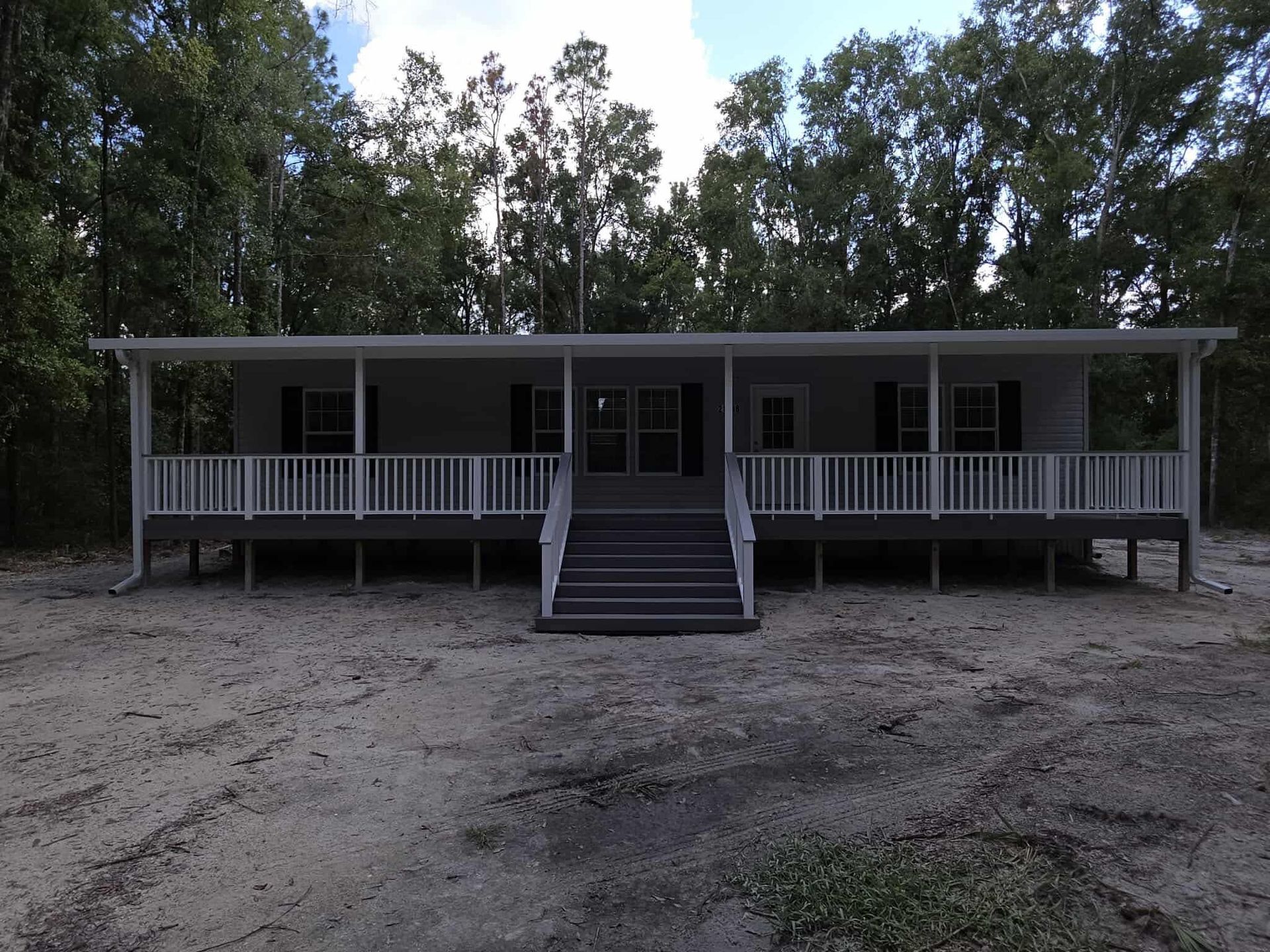 Small gray cabin with a central staircase and front porch, set among trees on a dirt clearing.