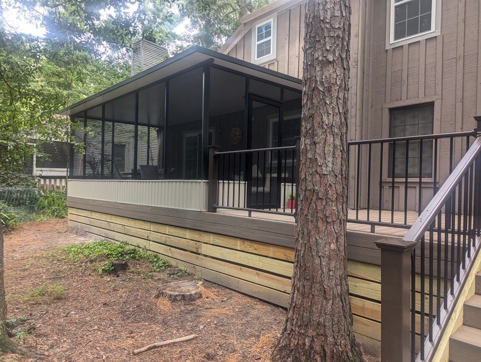 Screened porch with black railing and stairs on a house beside a large tree and wooded yard