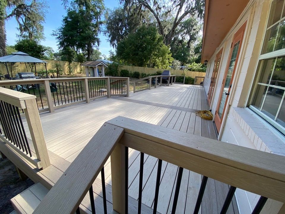 Wooden deck with railings beside a house, overlooking a pool and trees in bright sunlight