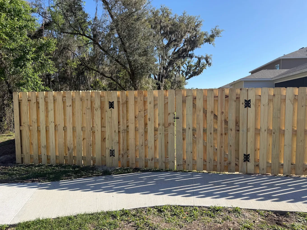 Wooden privacy fence along a sidewalk with trees and a house behind it