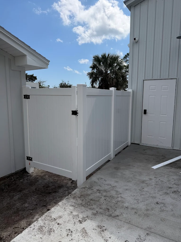 White privacy fence with gate beside a gray house and concrete patio under a blue sky.