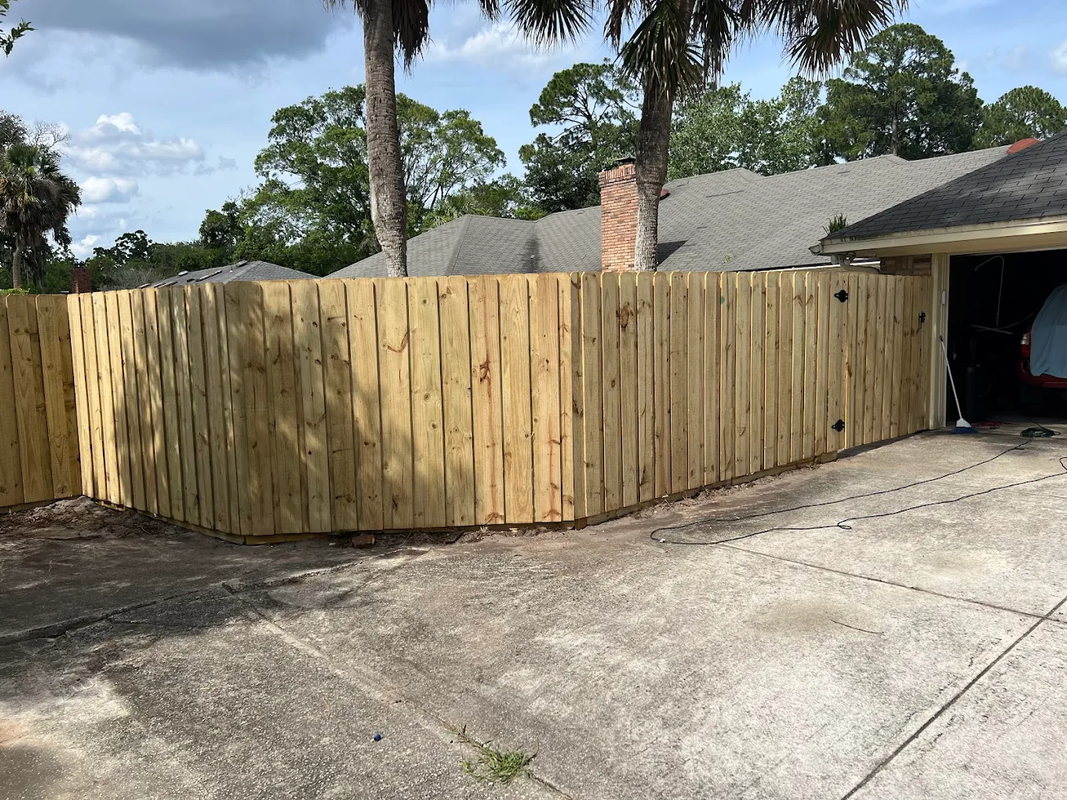 Wooden privacy fence enclosing a driveway beside a house, with trees and cloudy sky in the background