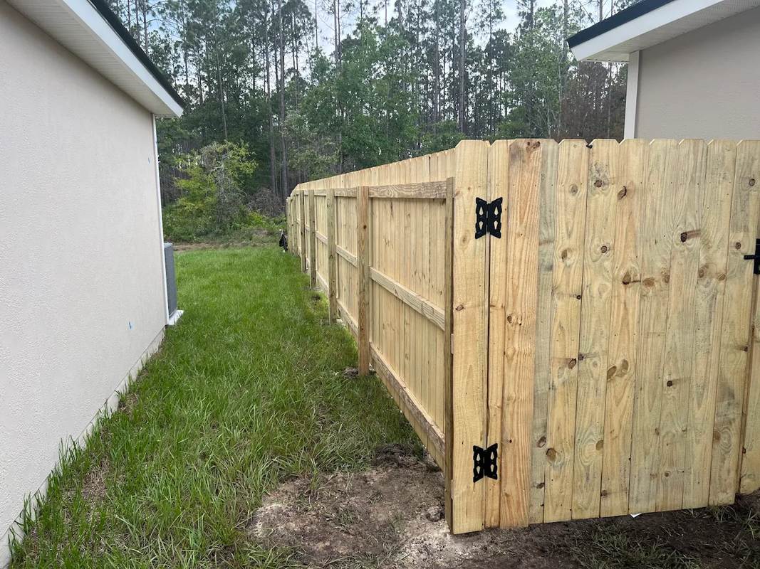 Wooden fence and open gate beside a white house, with a grassy yard and trees beyond.