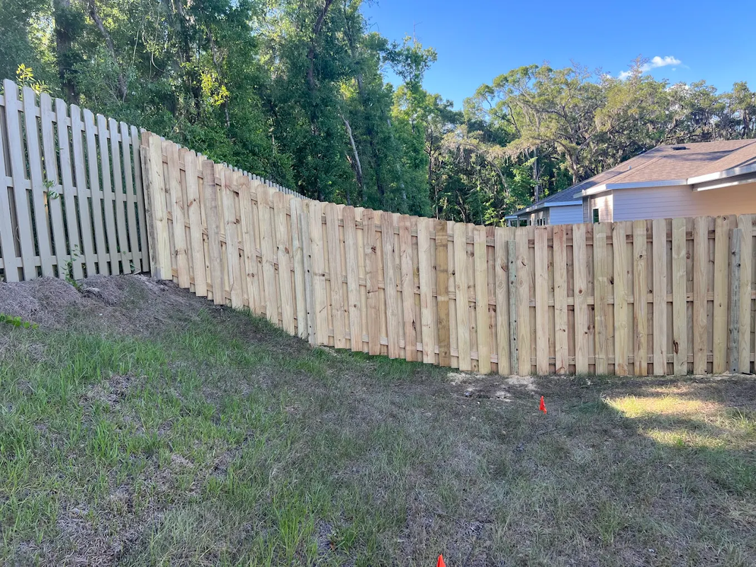 Curved wooden fence on a grassy sloped yard with trees and a house in the background