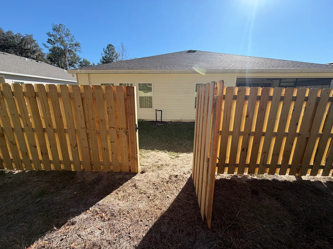 Open wooden gate leading to a sunlit backyard and a beige house with a gray roof.