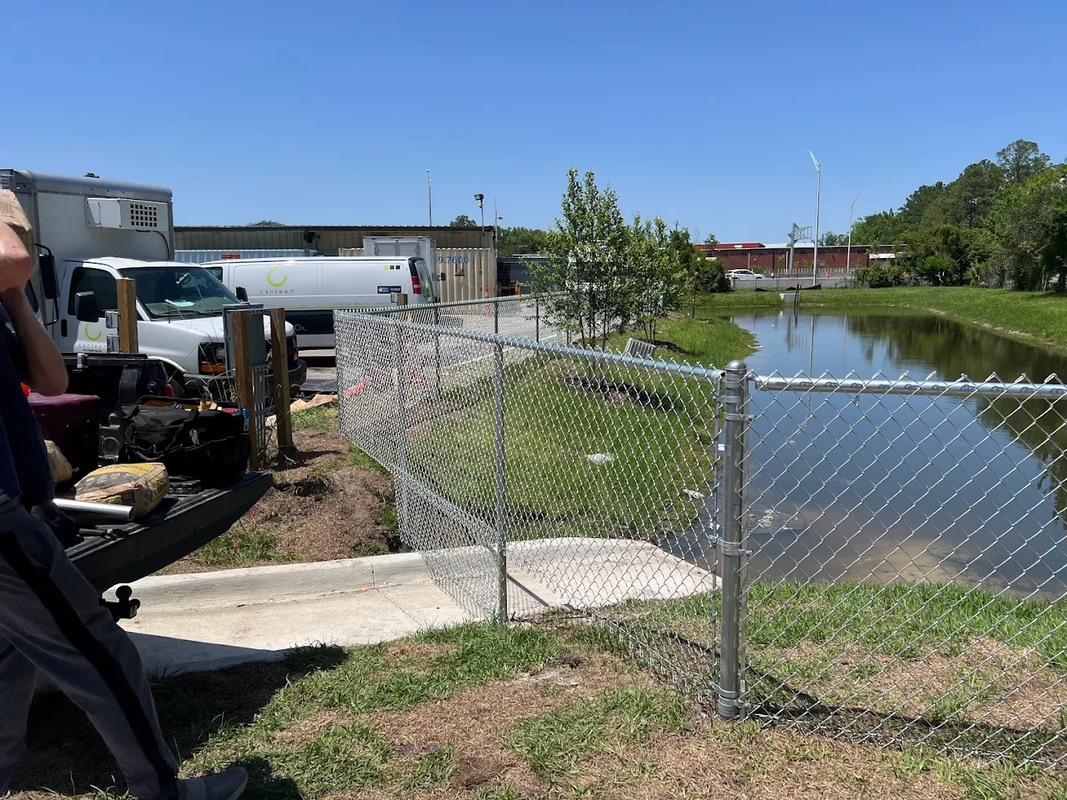 Person photographing a fenced pond beside an industrial site on a sunny day