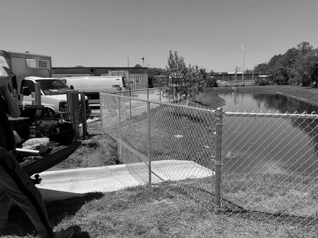 Chain-link fence beside a lake with workers and a concrete truck on the left, trees and buildings in the background