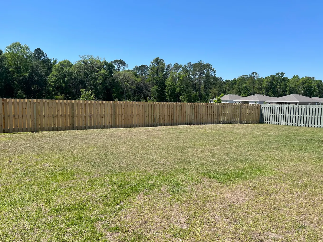 Wooden fence along a grassy backyard under a clear blue sky, with trees in the background.