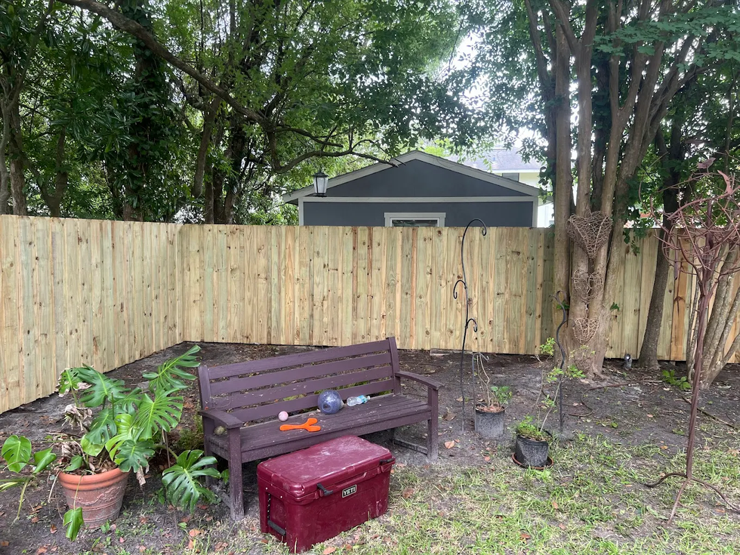 Backyard with wooden fence, small bench, red ottoman, potted plants, and trees in the background