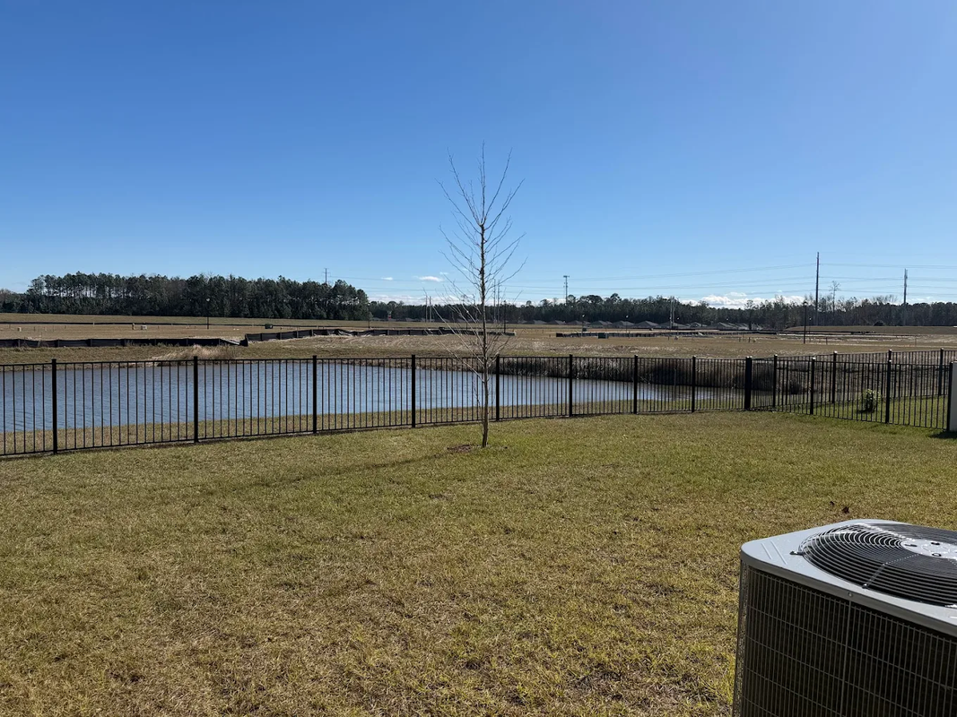 Grassy field beside a calm pond under a clear blue sky, with distant trees and utility posts.