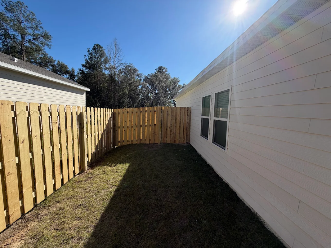 Narrow grassy side yard beside a white house and wooden fence under a bright blue sky.