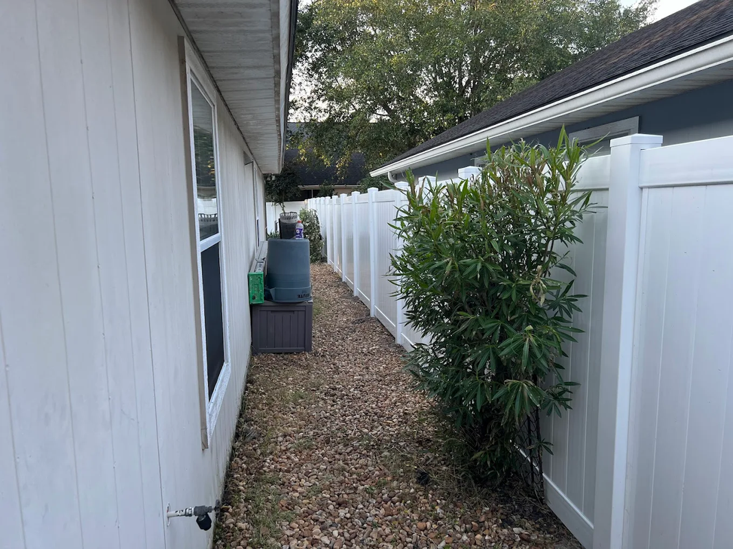 Narrow side yard with white fences, a house wall, and a leafy shrub beside a gravel path.
