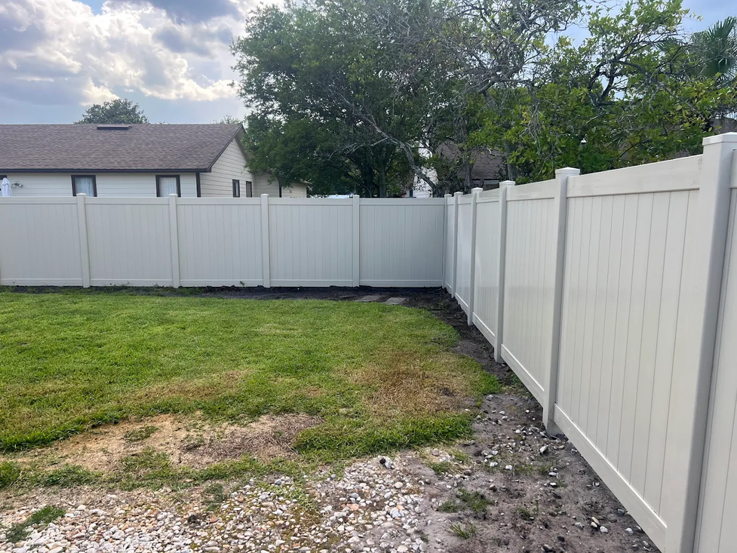 Backyard with white vinyl fence, patchy grass, and gravel beside a house and trees.