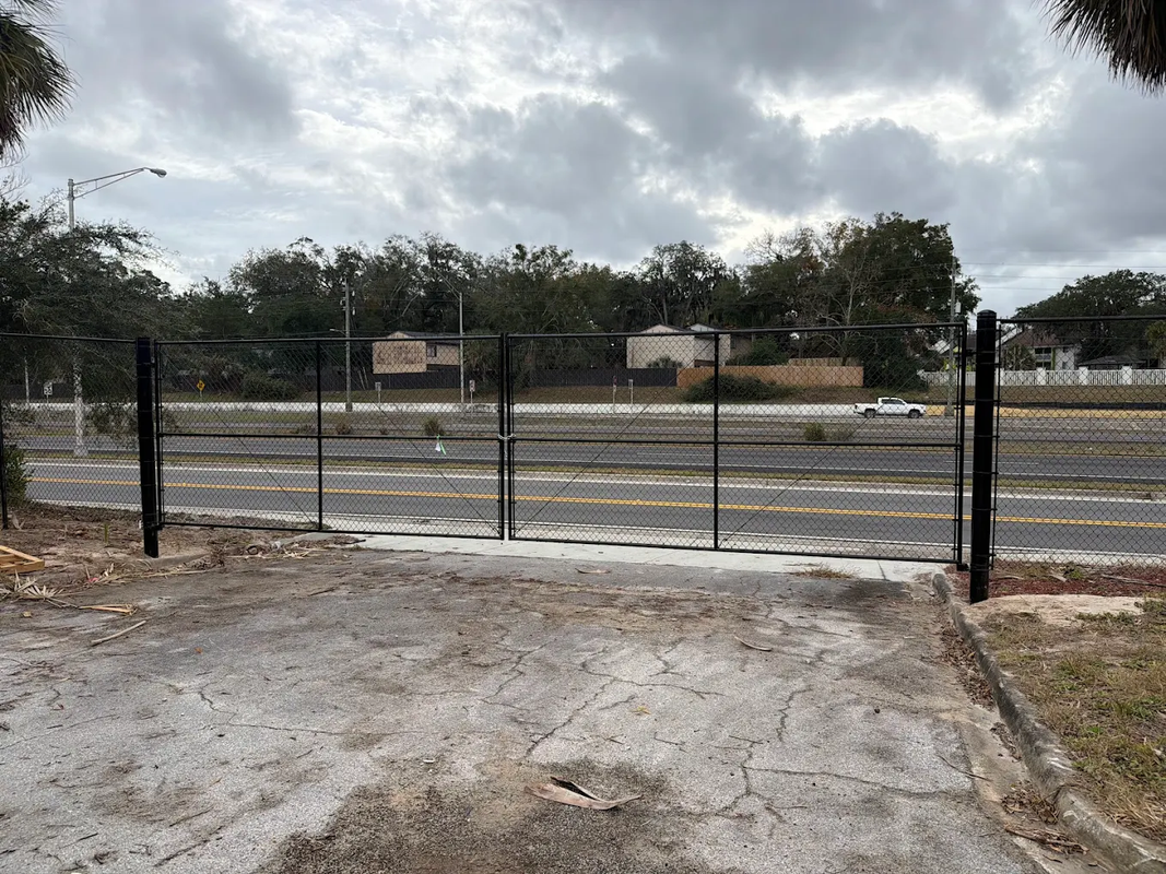 Chain-link gate across a gravel driveway under a cloudy sky, with trees and houses in the background