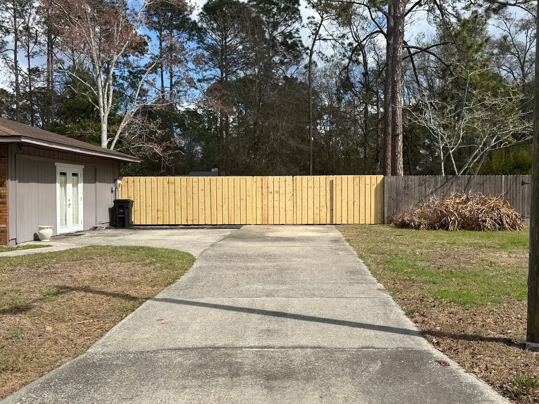 Concrete driveway leading to a tan wooden fence, with a house on the left and trees behind it.