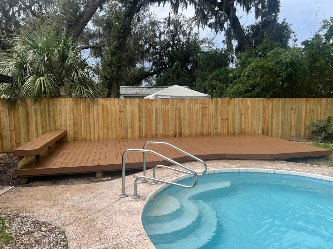 Backyard pool with wooden deck, metal handrail, and bamboo fence in front of trees