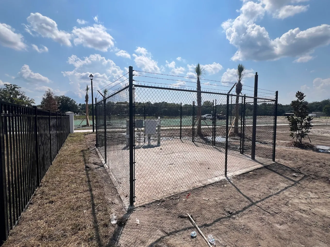 Chain-link fenced dirt court under a bright blue sky with clouds.