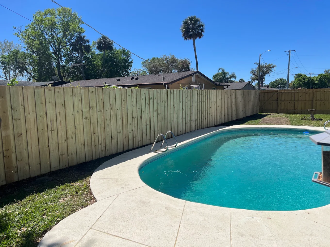 Backyard swimming pool with a wooden fence, clear blue water, and bright sunny sky