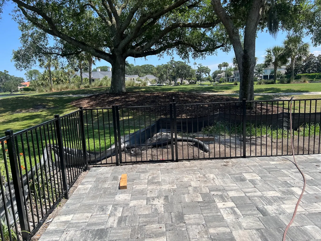 Stone patio overlooking a black metal fence, grassy park, and large trees under a bright blue sky.