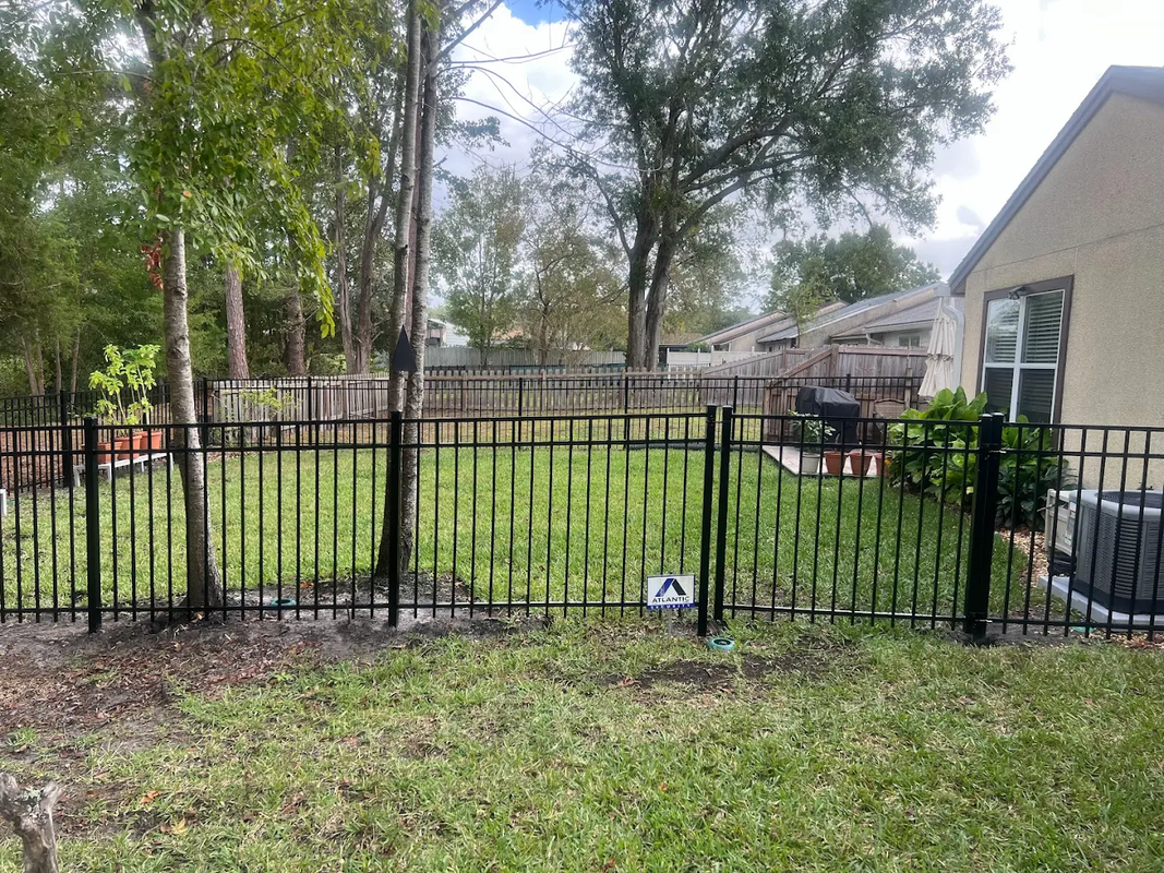 Backyard with black metal fence, green grass, trees, and a house beside a small soccer ball
