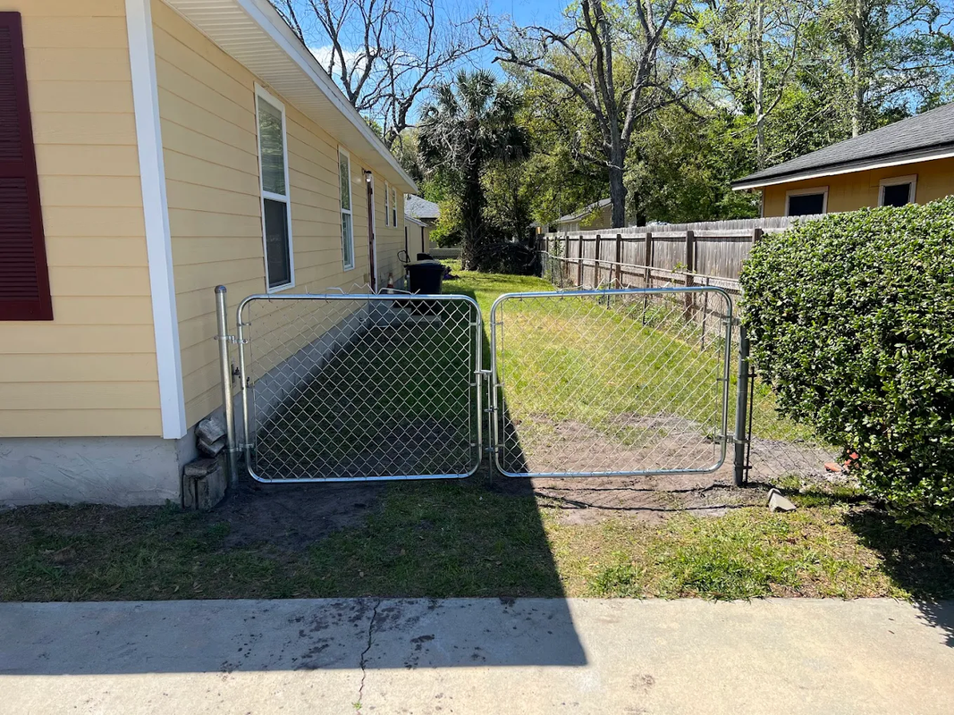 Backyard side yard with a yellow house, chain-link gate, fence, grass, and trees in sunlight