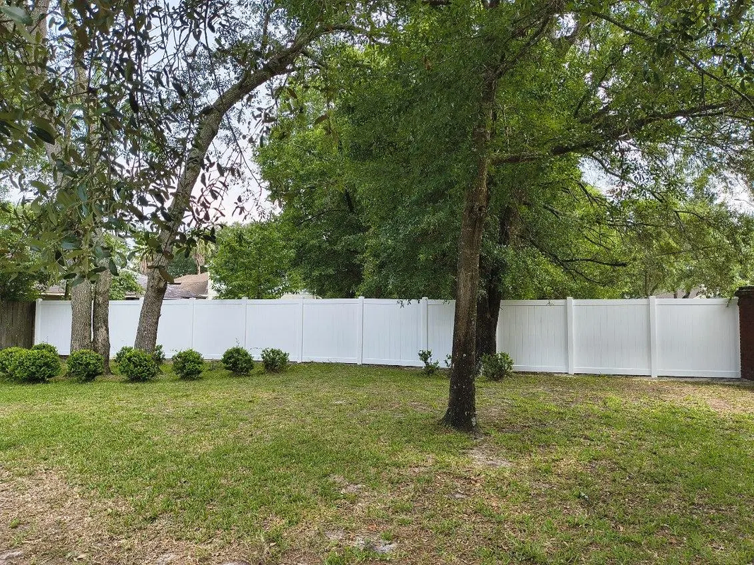 Grassy backyard with a large tree and a white privacy fence under cloudy skies