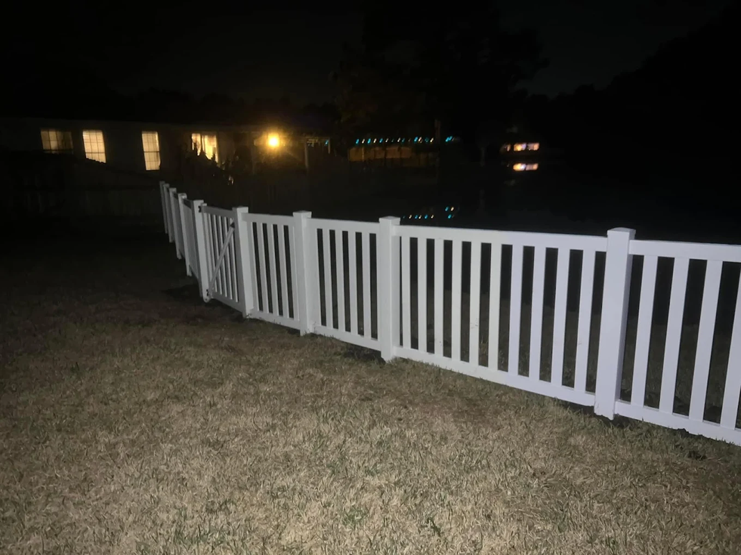 White fence lining a grassy yard at night, lit by a camera flash with house lights in the background