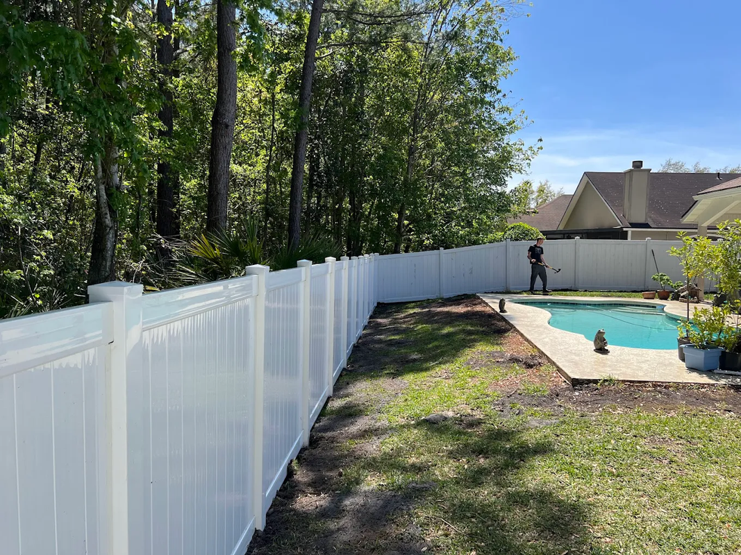 White vinyl fence beside a backyard pool, with trees lining the yard and a house in the background.