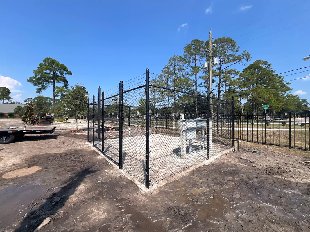 Black metal fenced enclosure with a small structure in a sandy outdoor area under a clear blue sky
