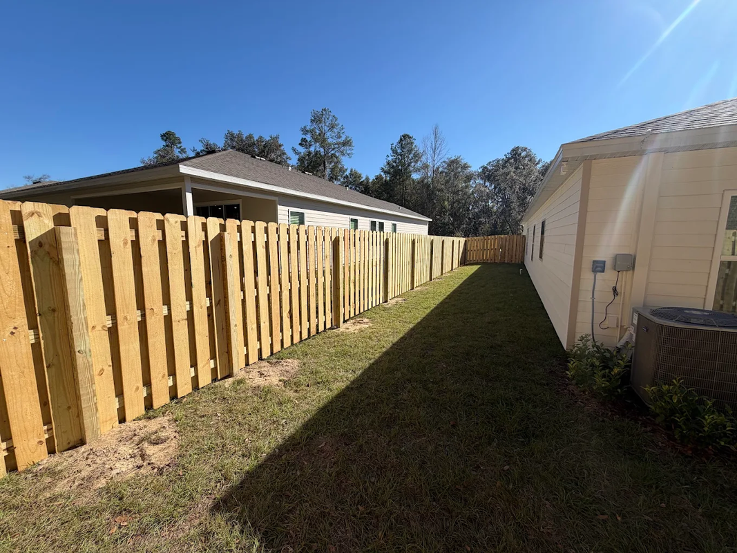 Narrow grassy side yard between a wooden fence and a house, under a clear blue sky.