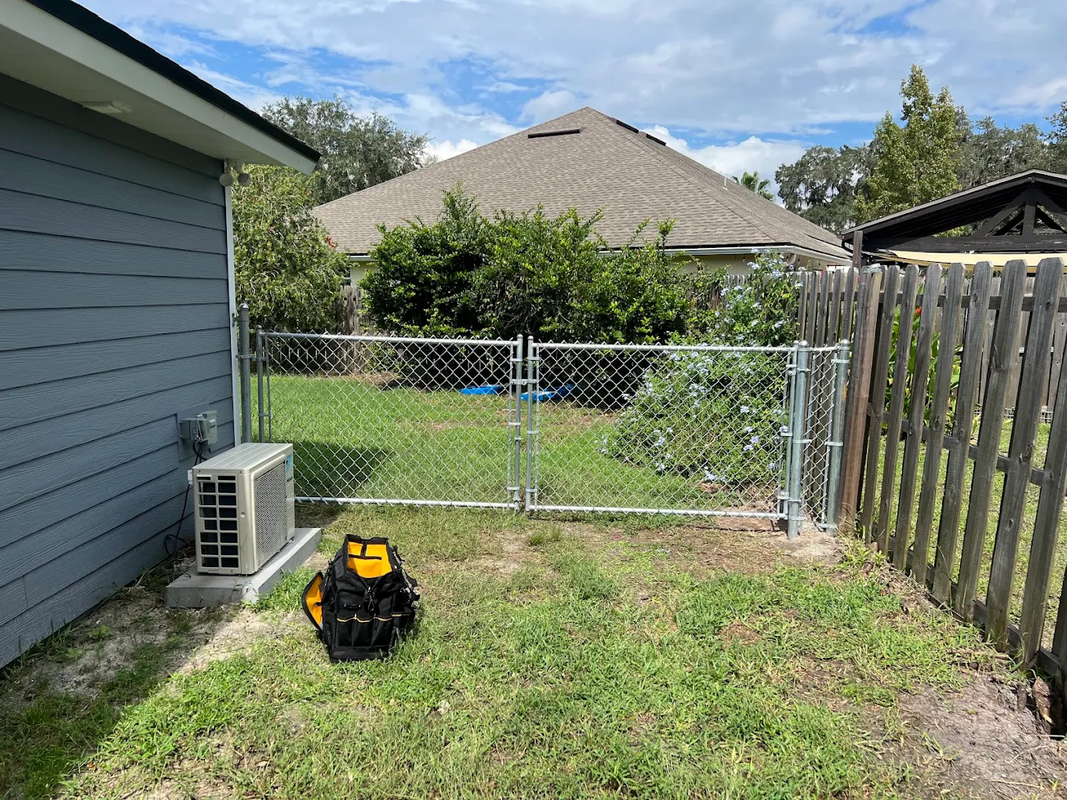 Backyard with chain-link gate, house siding, wooden fence, grass, and a lawn mower in the foreground