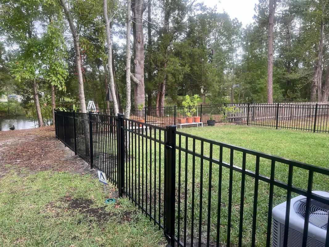 Black metal fence along a grassy yard with tall trees and a pond in the background