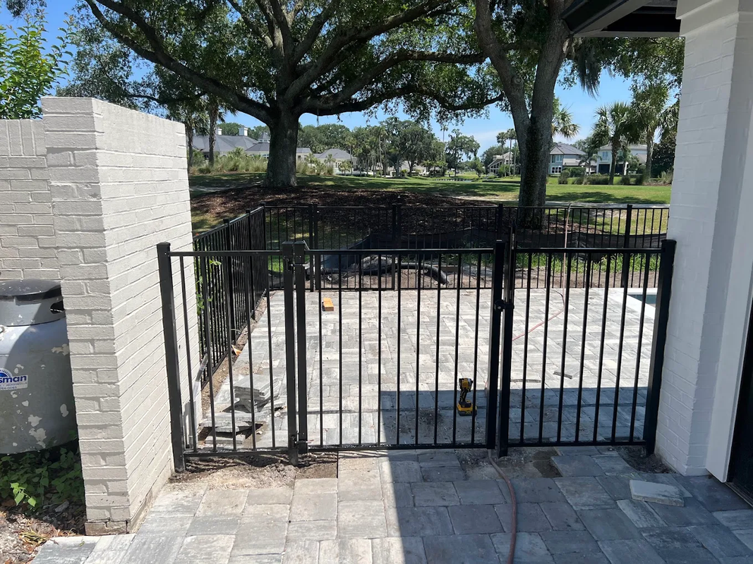 Black metal gate across a stone driveway beside a carport, with trees and a fenced yard beyond.