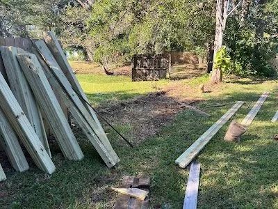 Stacked white boards outdoors beside scattered lumber in a grassy yard with trees and a small shed.