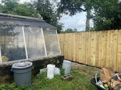 Backyard garden with a greenhouse, trash bins, wooden fence, and cut logs on grass