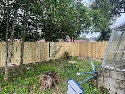 Backyard with a wooden fence, trees, lawn, and a tipped blue chair near a greenhouse structure