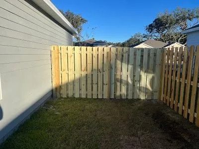 Small fenced backyard with grass, a white house wall on the left, and blue sky above.