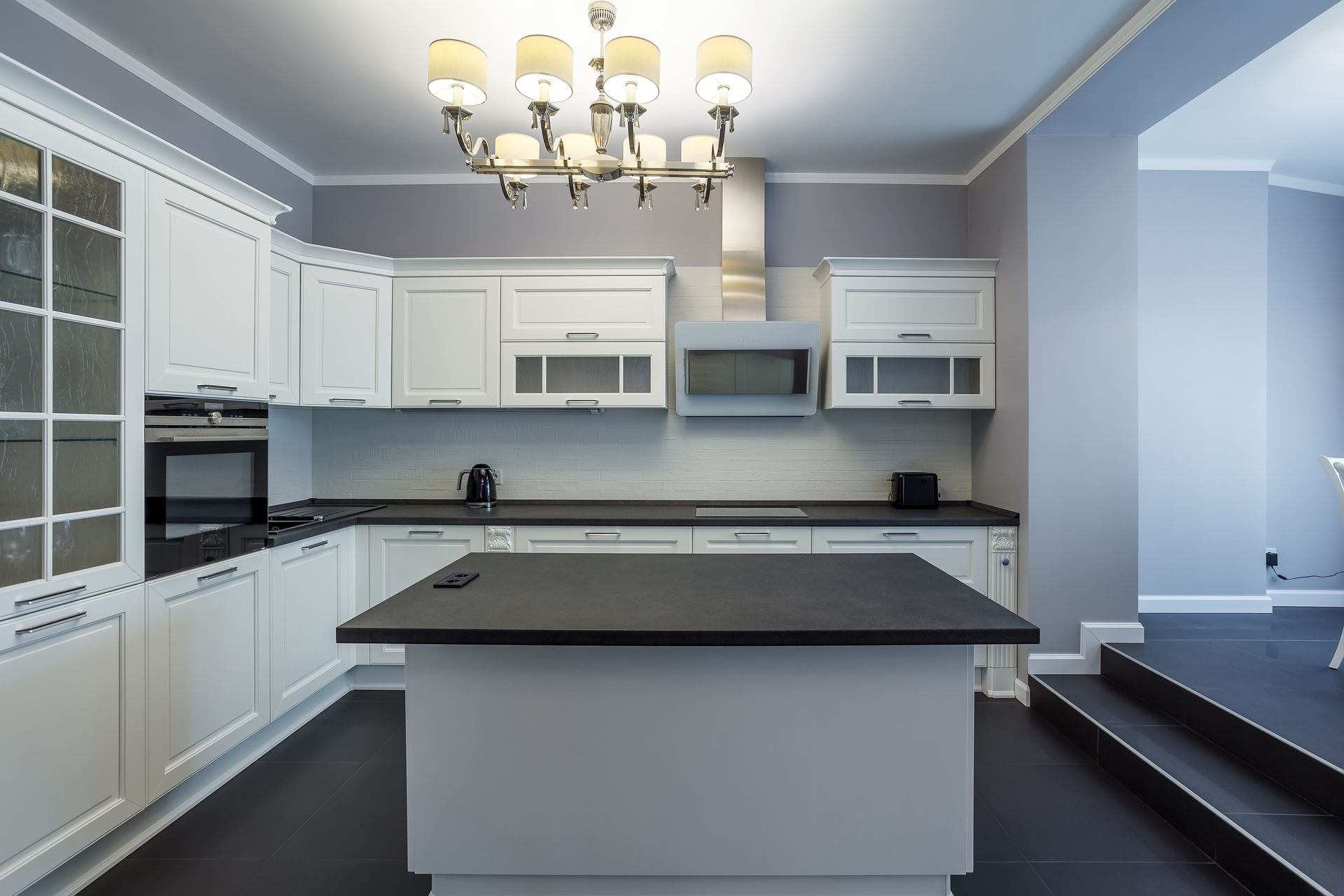 White kitchen with black countertops, island, and appliances. Chandelier hangs above. White kitchen with black countertops, island, and appliances. Chandelier hangs above.