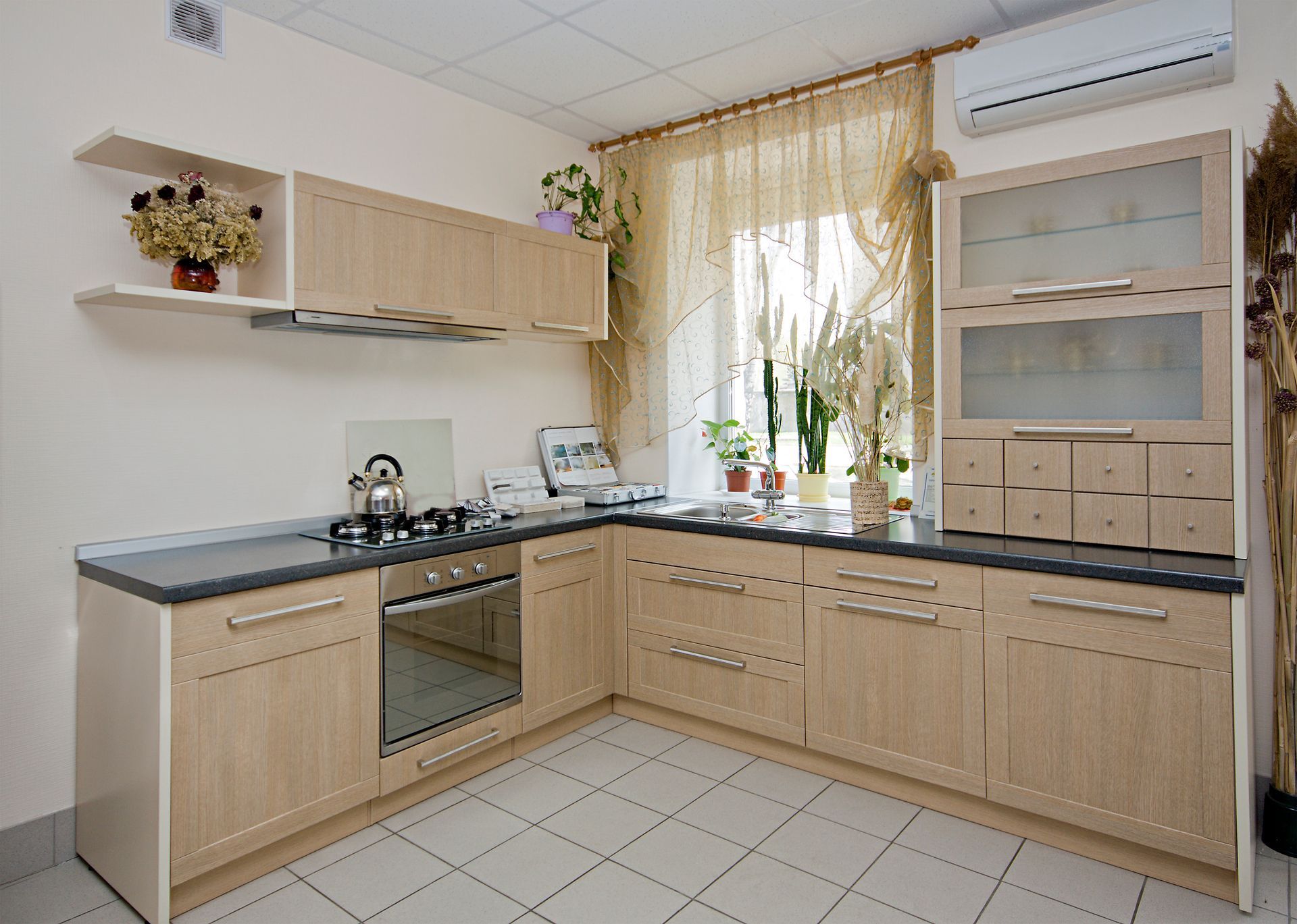 L-shaped kitchen with light wood cabinets, gray countertops, and a window with sheer curtains. L-shaped kitchen with light wood cabinets, gray countertops, and a window with sheer curtains.