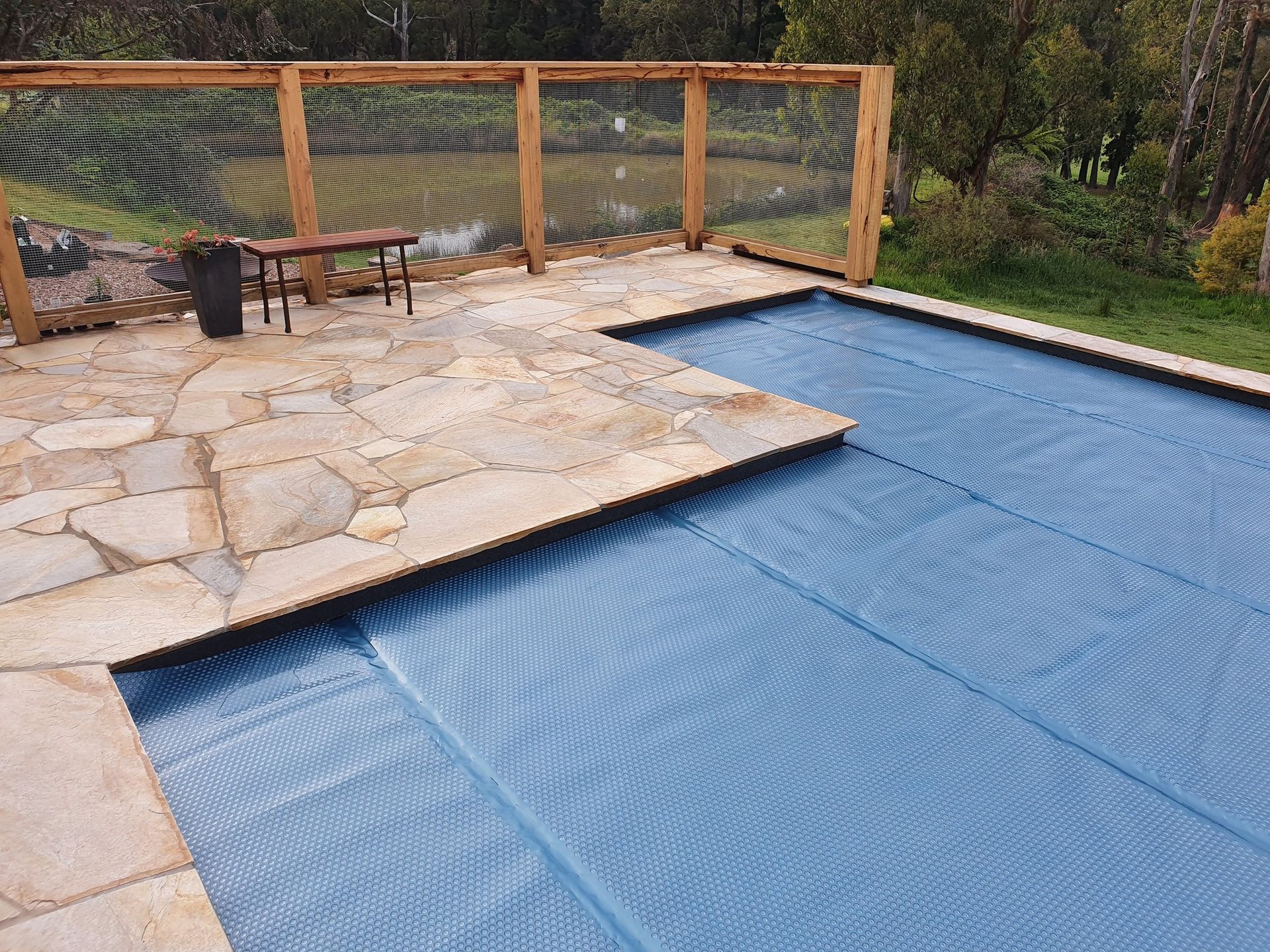 Pool covered with blue tarp, surrounded by stone patio, wooden fence, and green grass.