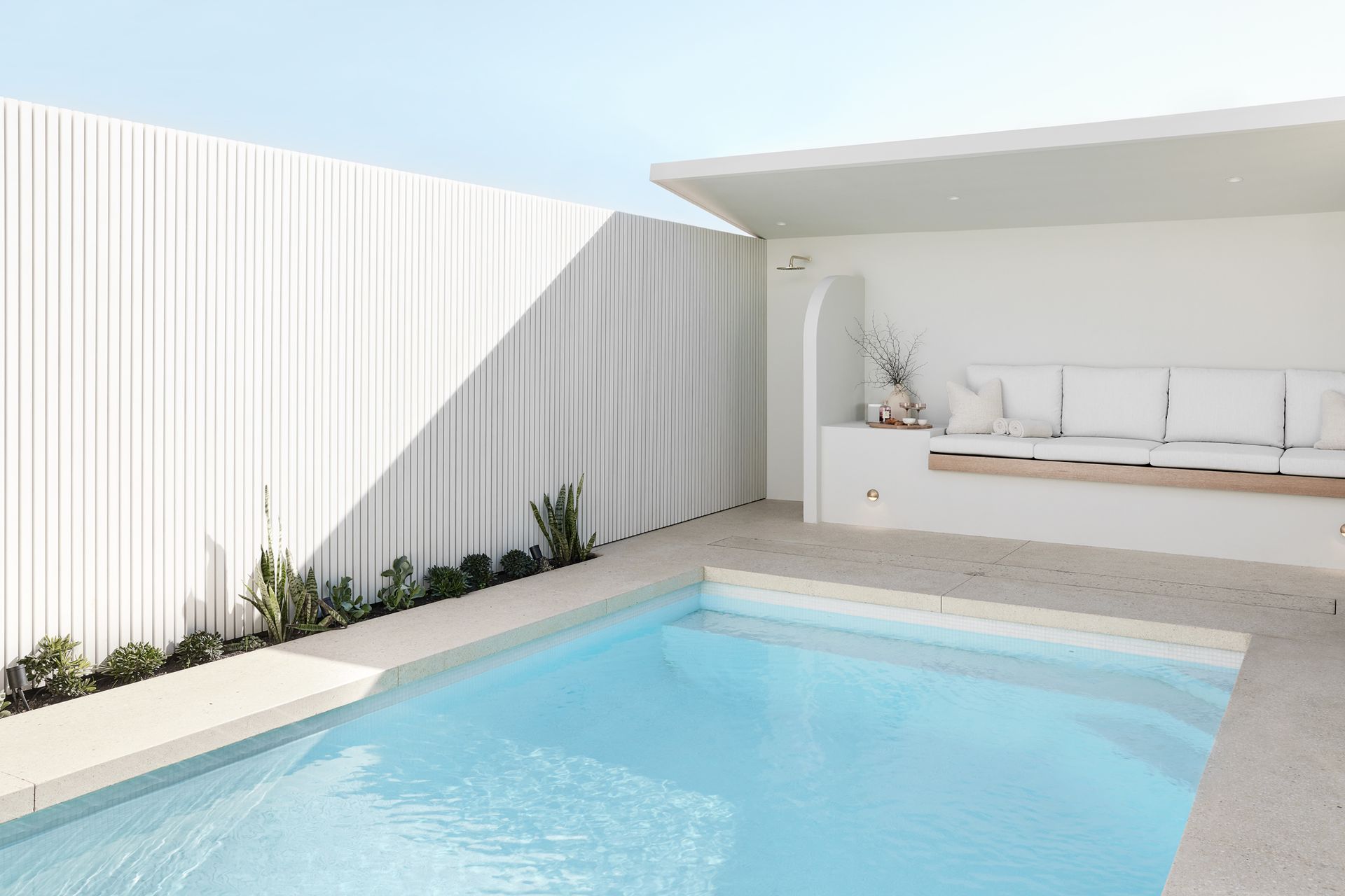 Rectangular blue swimming pool surrounded by stone tiles and a green hedge under a bright sky.