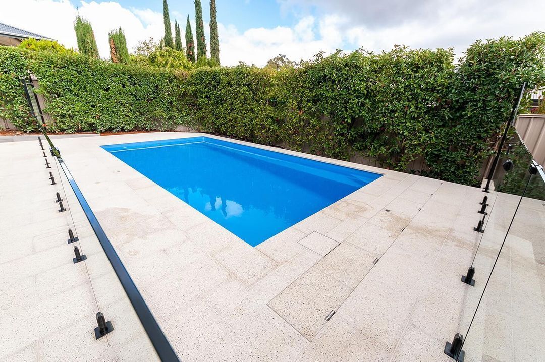 Blue rectangular pool with white stone patio surrounded by green hedges.